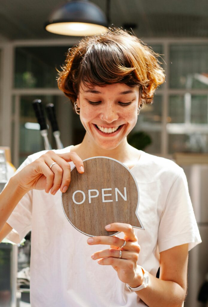 Happy female worker holding wooden Open sign in modern cafe, showcasing the impact of digital marketing for small business.